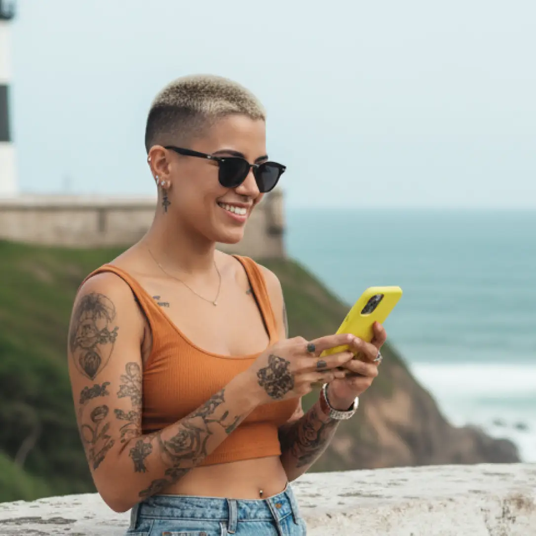 Mulher sorridente com cabelo raspado, tatuagens visíveis e óculos escuros, usando um celular amarelo em ambiente litorâneo. Representa liberdade e facilidade de usar o Jota para gerenciar finanças de qualquer lugar.